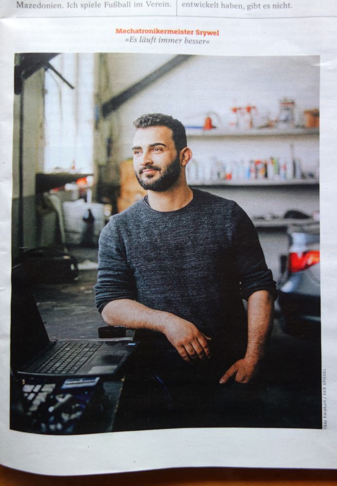 Imad Srywel, der sich lächelnd an die Werkbank lehnt, mit dem rechten Ellbogen auf diese gestützt. Er blickt nach links, von wo das Licht einfällt, in eine unbestimmte Ferne - vielleicht aus einem Fenster und vermutlich auch optimistisch in die eigene Zukunft. 
Quelle der Abbildung: ARP, Susmita; ELGER, Katrin; HAIDAR, Asia; OLBRISCH,Miriam: Ziemlich geschafft; DER SPIEGEL Nr. 32 / 1.8.2025, S. 8-15, S. 13.