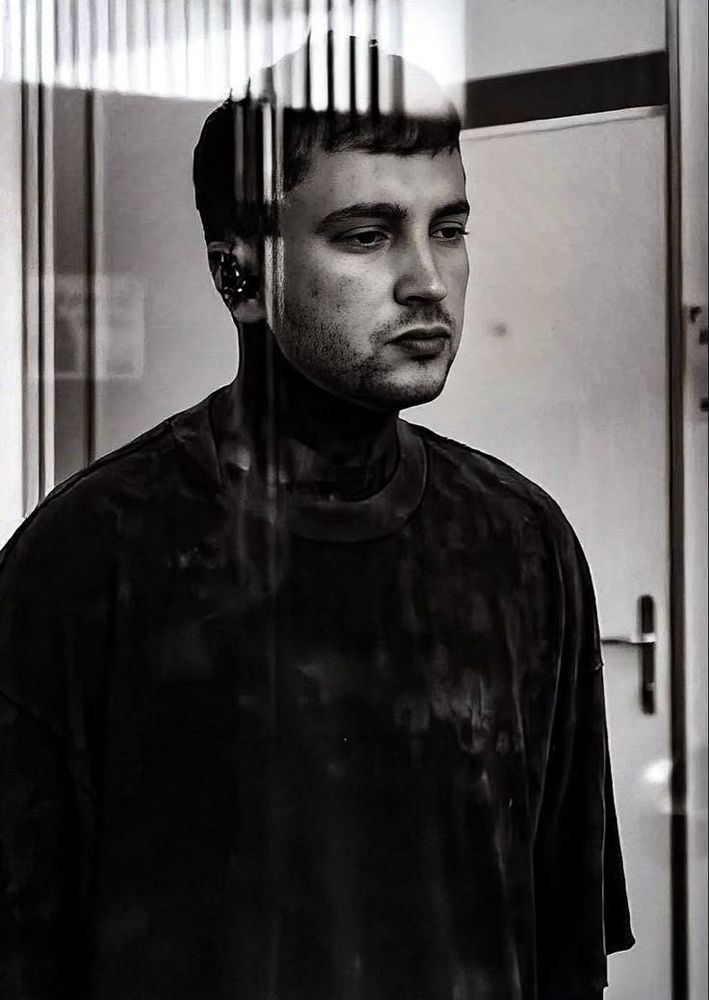 Black and white photo of Tyler looking through a window. He looks very deep in thoughts and/or disassociated. 