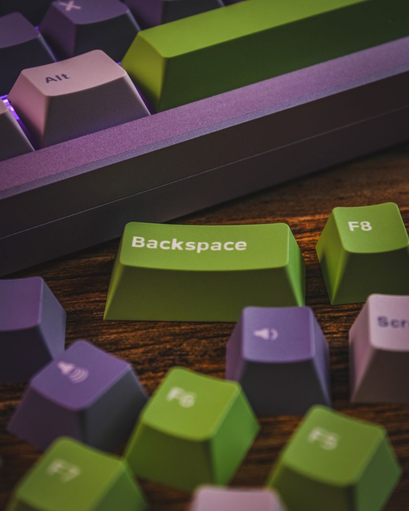 Close-up of a mechanical keyboard with green, purple, and lilac keycaps. The green "Backspace" key rests on a wooden surface, surrounded by loose keys like "F6" and "F8." 