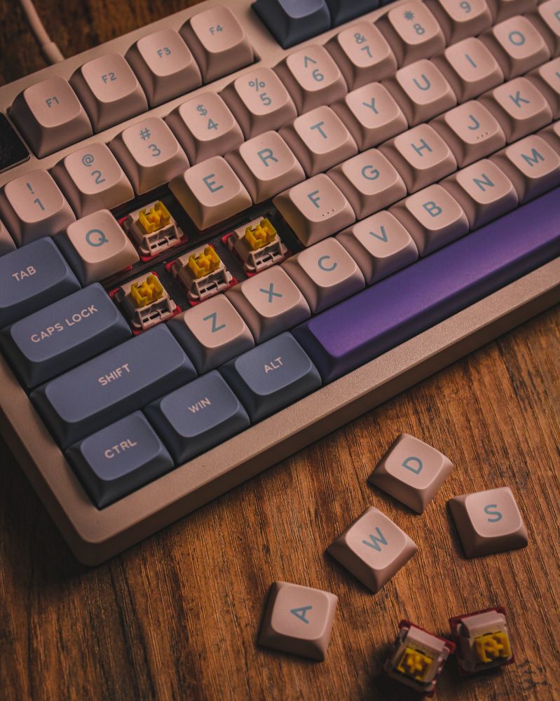 A LEOBOG Hi75 mechanical keyboard with pastel-colored keycaps in shades of lavender, beige, and blue, displayed on a wooden surface. Several keycaps have been removed to reveal yellow tactile switches underneath, while the "AWDS" keys and a few switches are scattered nearby. The keyboard's design and warm lighting emphasize its sleek and modern aesthetic.