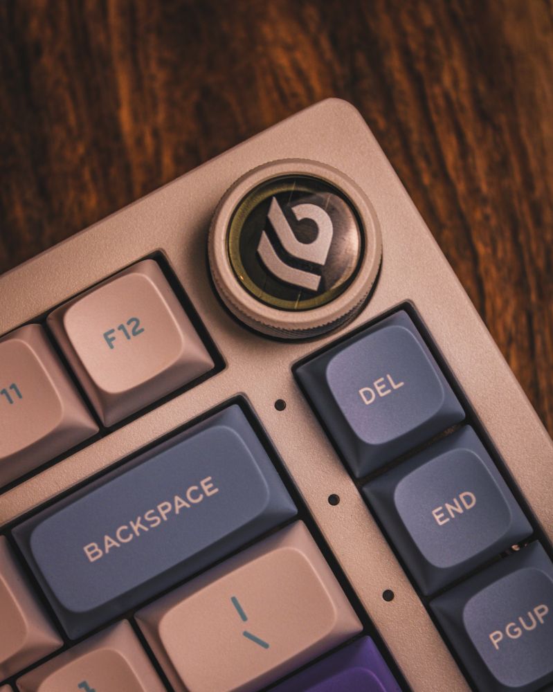 A close-up view of the LEOBOG Hi75 mechanical keyboard, highlighting the top-right corner. The design features a metal knob with a custom insert displaying a stylized "B" logo in white against a black background. Surrounding keys, such as "BACKSPACE," "DEL," "END," and "PGUP," have pastel-colored keycaps in lavender, beige, and blue, complementing the keyboard's sleek and modern aesthetic. The wooden surface beneath adds warmth to the composition.
