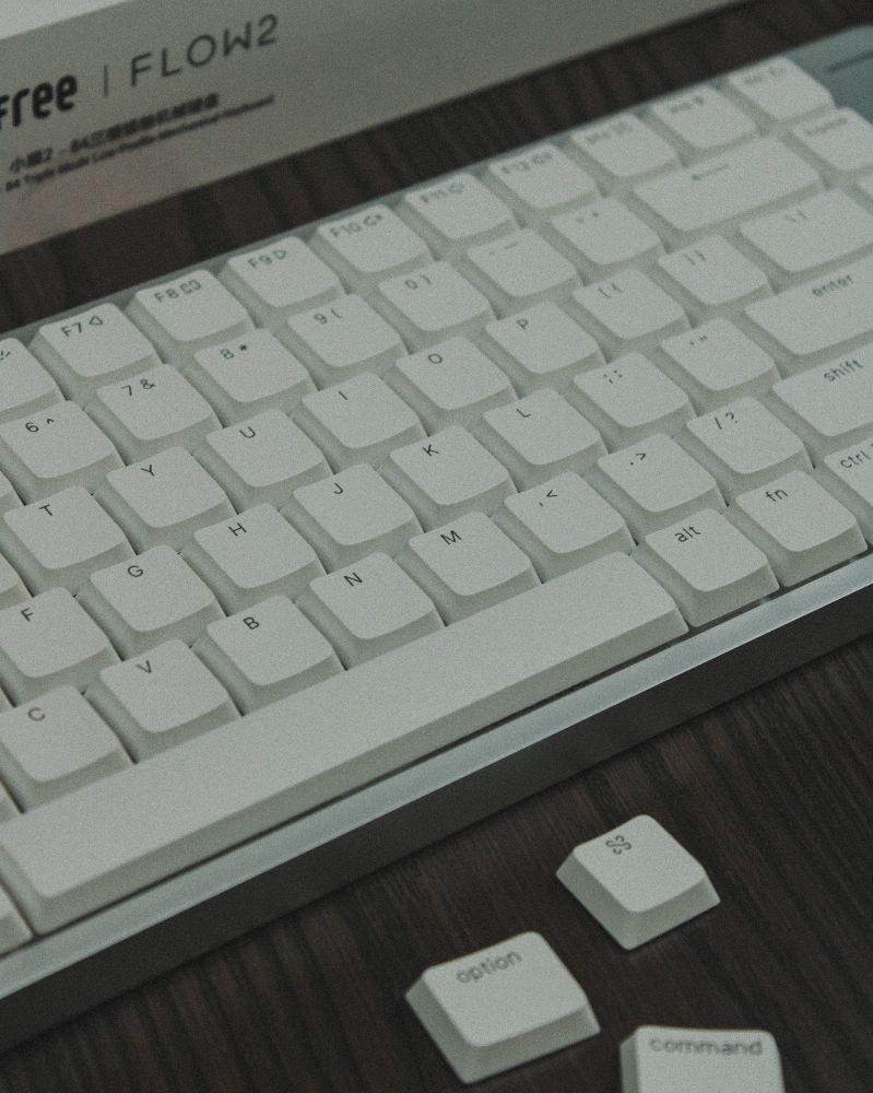 Silver low profile keyboard with white keycaps on a wooden desk. The mac os specific keycaps sit below it in shot. 