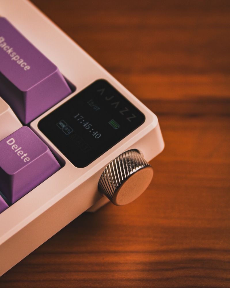 A close-up of a white mechanical keyboard with a metallic rotary knob on the top right corner. Next to the knob, there is a small screen displaying the brand "Ajazz," along with the date, time, and battery level. The surrounding keycaps are lavender and white, adding a vibrant touch to the design. The keyboard rests on a wooden surface.