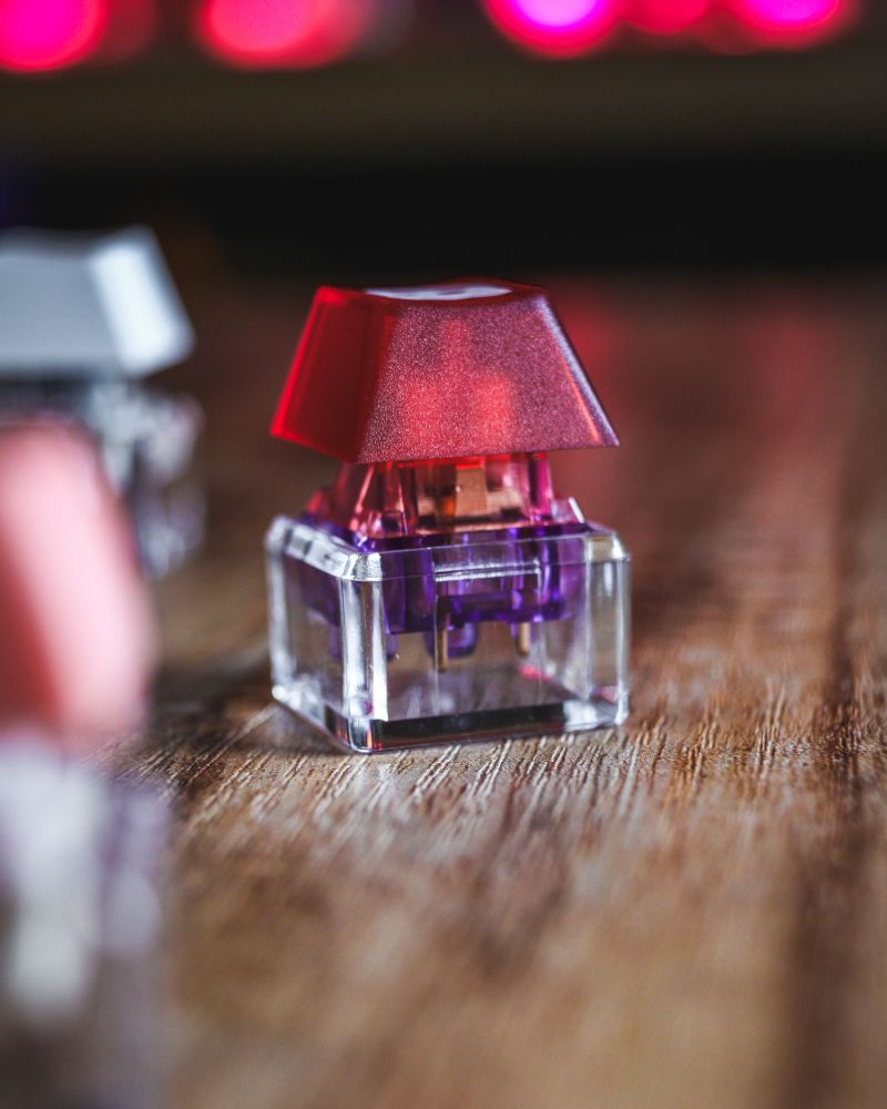 Close-up of a red keycap on a transparent mechanical switch with a purple stem, set against a wooden surface and soft bokeh lights in the background.