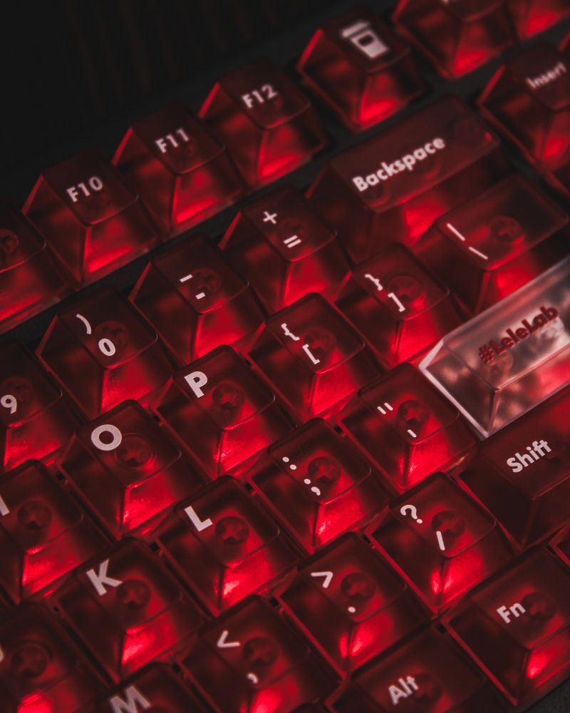 A close-up of a mechanical keyboard featuring translucent red keycaps illuminated by a vibrant red backlight. The keycaps have clear legends, including function keys like "Backspace," "Shift," and special characters. A branded "#leleLab" keycap is visible, adding a unique touch to the design.