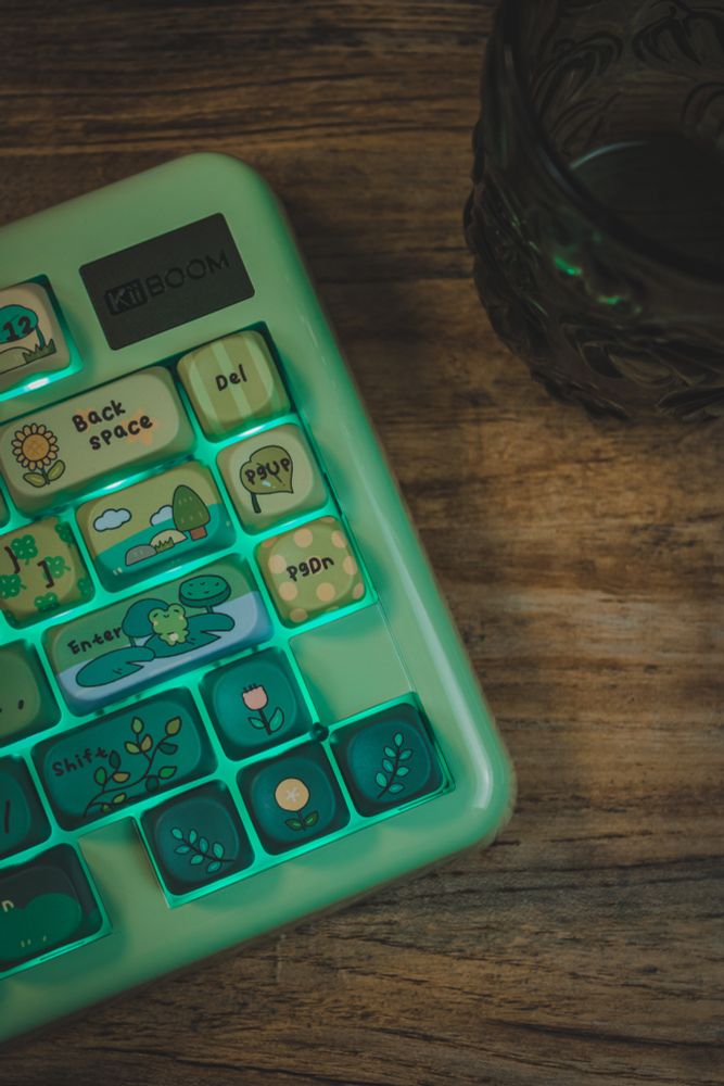 Close-up of a pastel green KiiBOOM keyboard with cute, nature-themed keycaps glowing softly on a wooden desk.