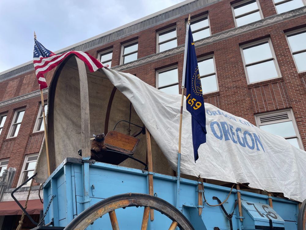 View of a light blue historic-looking wagon with an Oregon State flag and a National flag attached to it. In the distance is a red-brick building with lots of windows. A stuffed bison toy is sitting on the bench of the wagon.