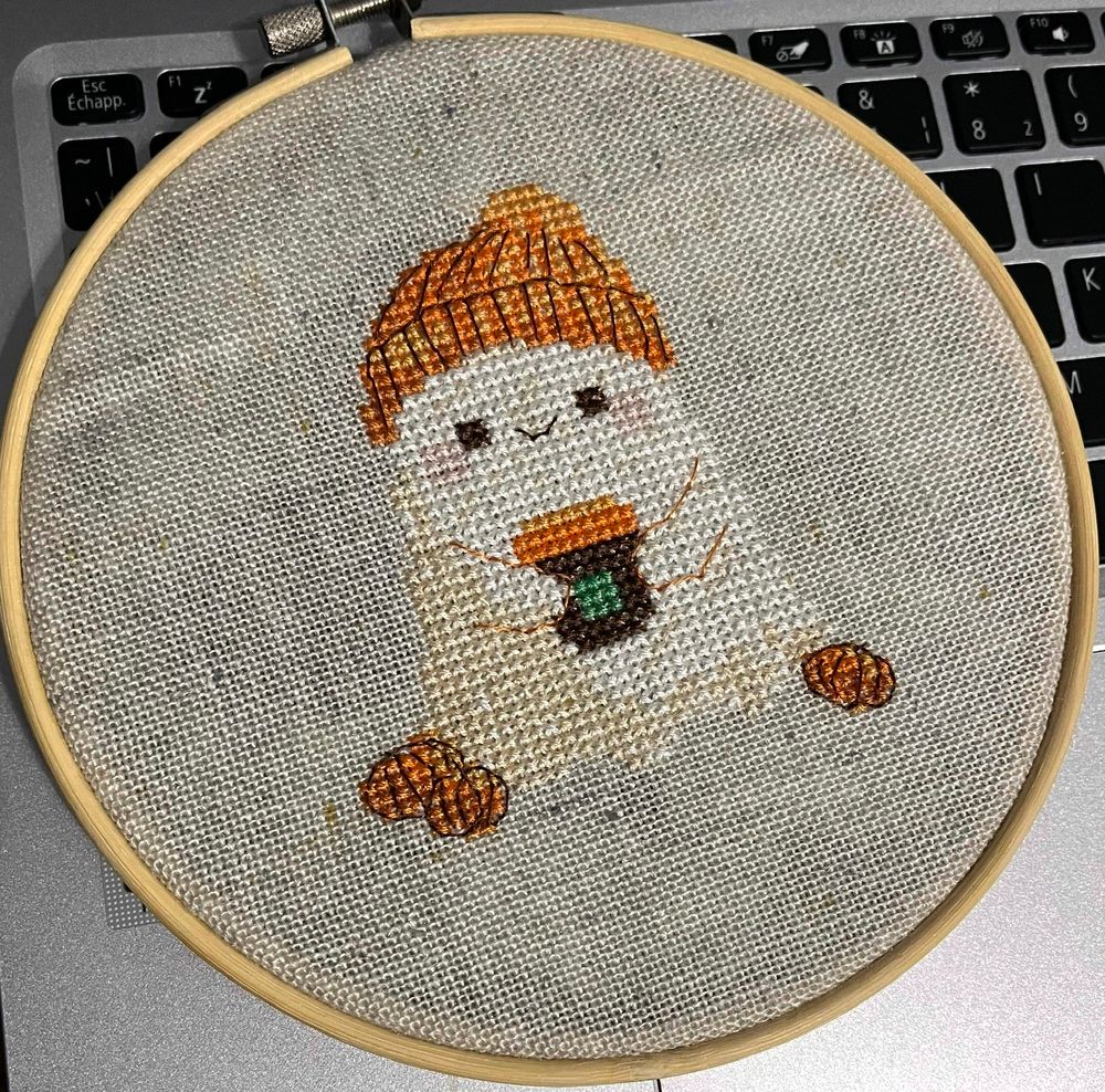a cross stitch of a what ghost wearing an orange toque and holding a cup of coffee on grey-ish linen fabric. 