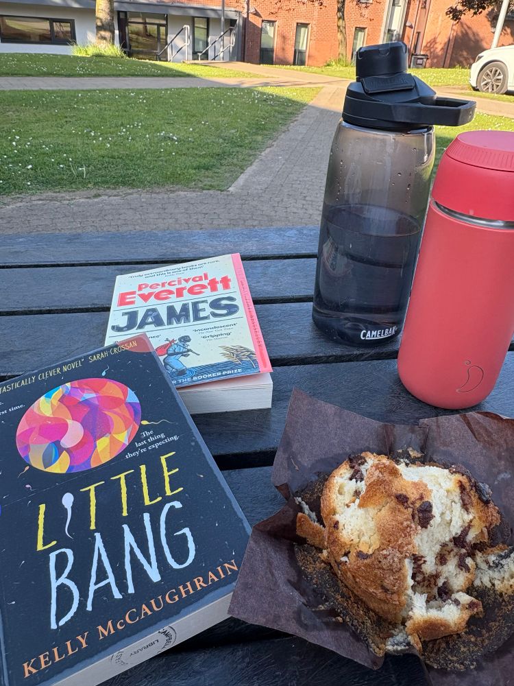 A photograph taken outside on a sunny day. Two books are on a picnic bench next to a muffin, a travel cup of coffee, and a bottle of water. The books are ‘James’ by Percival Everett and ‘Little Bang’ by Kelly McCaughrain.