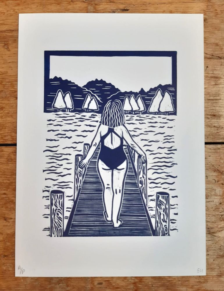 A lino print of a wild swimmer preparing to take a dip. The swimmer is walking along a wooden platform. The water is calm and in the distance, there are trees and mountains. Ink colour is navy blue. 