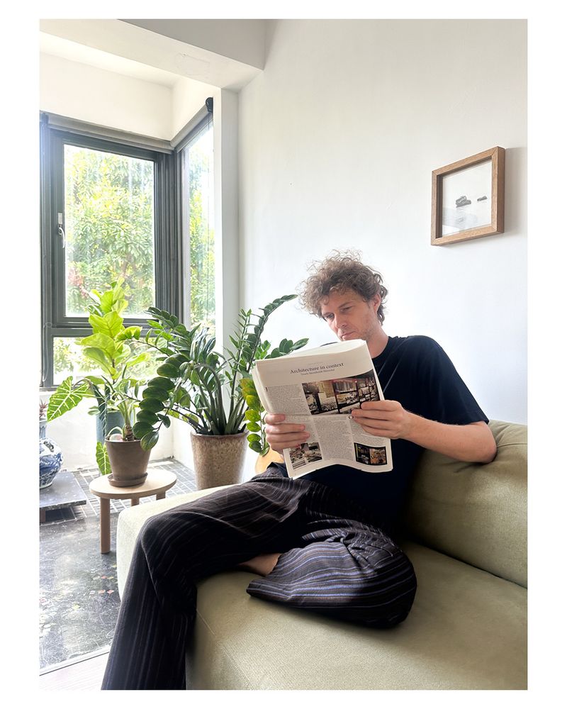 A white man sitting on a light green couch, in dark pants and t-shirt reading a paper, behind him is a white wall and the his left pants in front of a window looking out onto a tree.