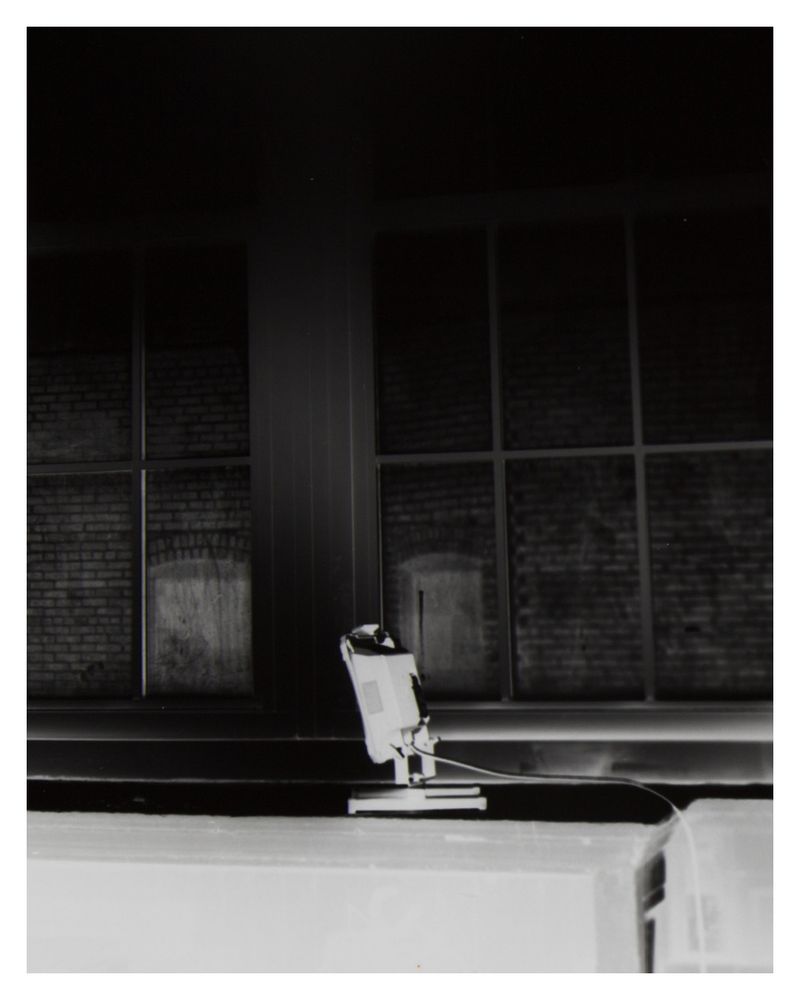 Inverted black and white silver gelatin, excerpt. Centered to the bottom is the back of a floodlight rendered light gray contrasted on the black background, standing on an open cabinet with pipes and a radiator visible.