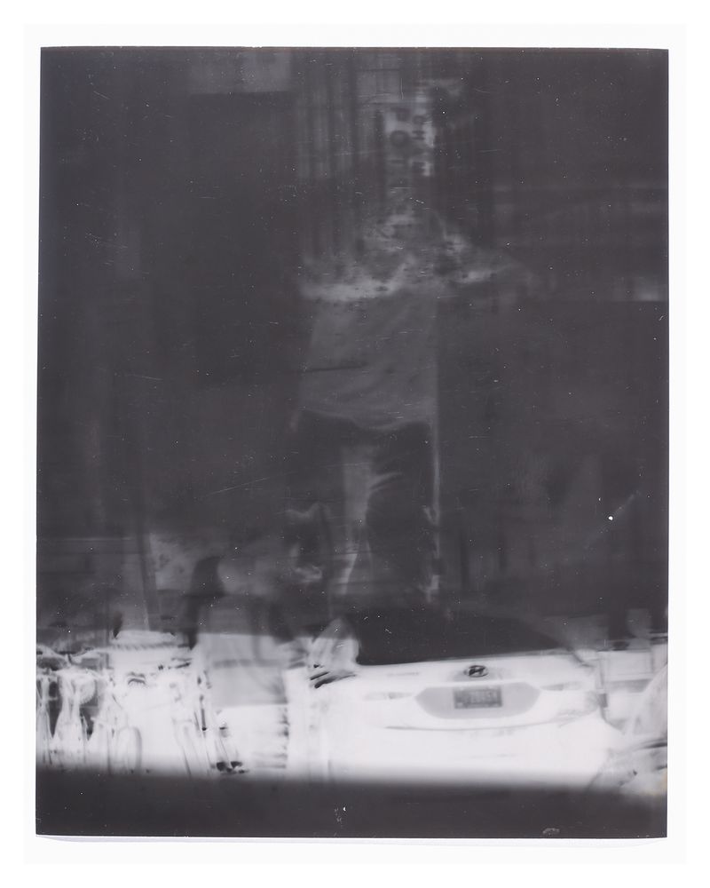 8x10 inch portrait inverted black and white silver gelatin; a dark composition is cut by a horizontal band of light across the bottom, in this are the rear end of a few cars visible, above them a full figure is standing, their head looks like it’s engulfed in flames.