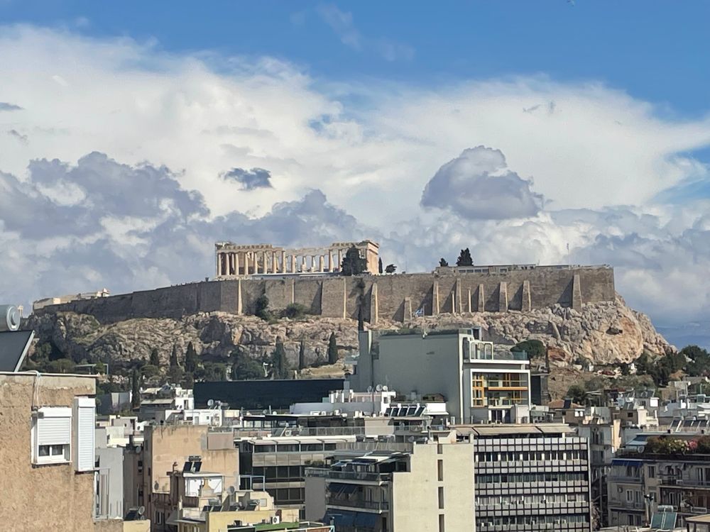View out a window over the rooftops towards the Athenian acropolis. The ruins of the Parthenon are highlighted and framed by a bright blue sky and fluffy clouds. 