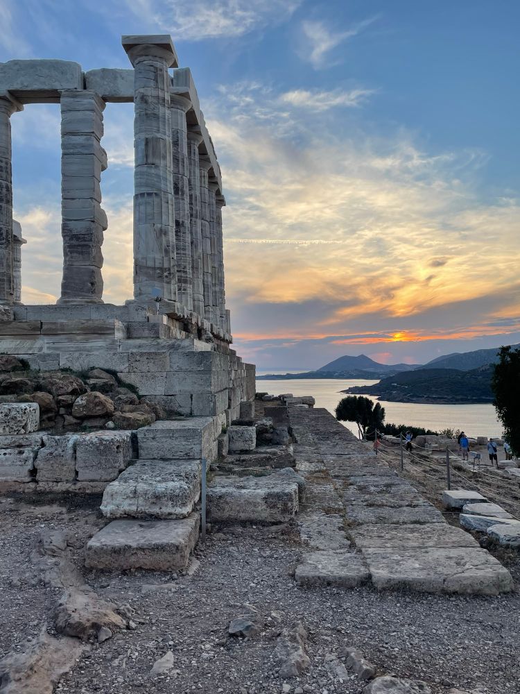 View across the ruins of the Temple of Poseidon (a platform and restored columns) to the Aegean Sea. The sea reflects the orange lines of the sky in muted tones.The sun is a tiny dot behind dark blue and yellow clouds on the horizon, with twilight’s bright blue sky overhead. 