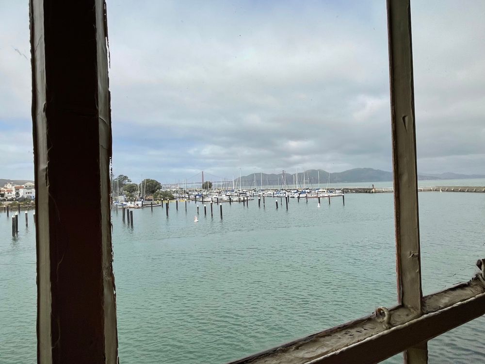 View out a window over a harbor, looking towards Golden Gate Bridge. The sky is grey, the water is a grey-green. There are boats moored in the distance and dark hills to the right, where the bridge enters Marin County.