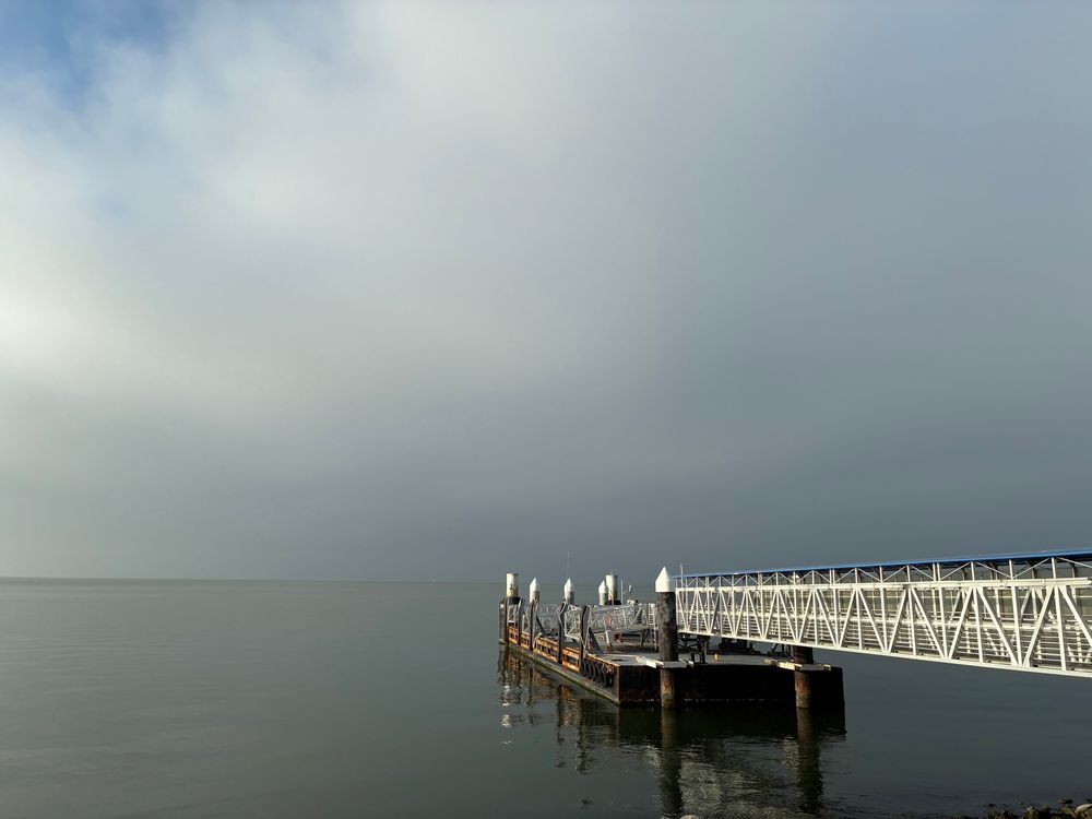 The water is a smooth sheet of grey. A pier stretches out into the water from the right of the photo; the upper two-thirds of the photo is blue-grey fog and clouds. In the upper left corner a patch of blue sky peeks through wisps of cloud. 