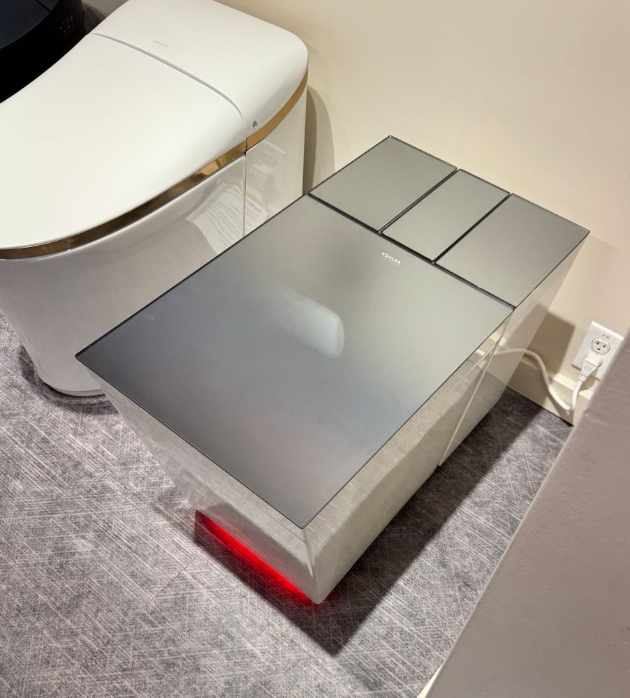 A boxy silver toilet that is shiny on top and has weird red lighting underneath. It’s plugged in to the wall.