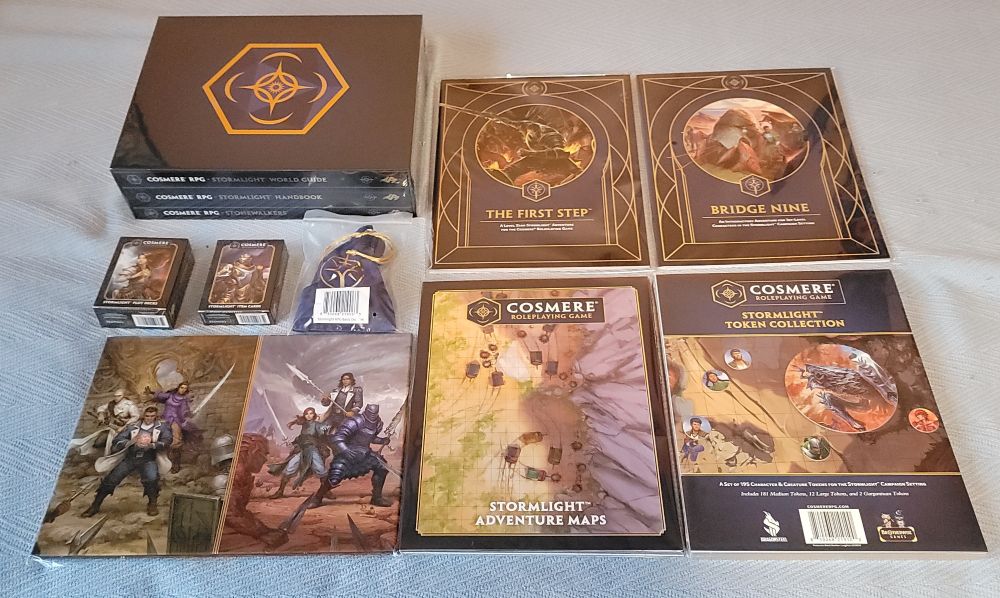 A photograph of the physical kickstarter rewards for the Cosmere RPG, including several Stormlight rulebooks, tokens and maps, a gm screen, two decks of cards, and a dice set in a bag.