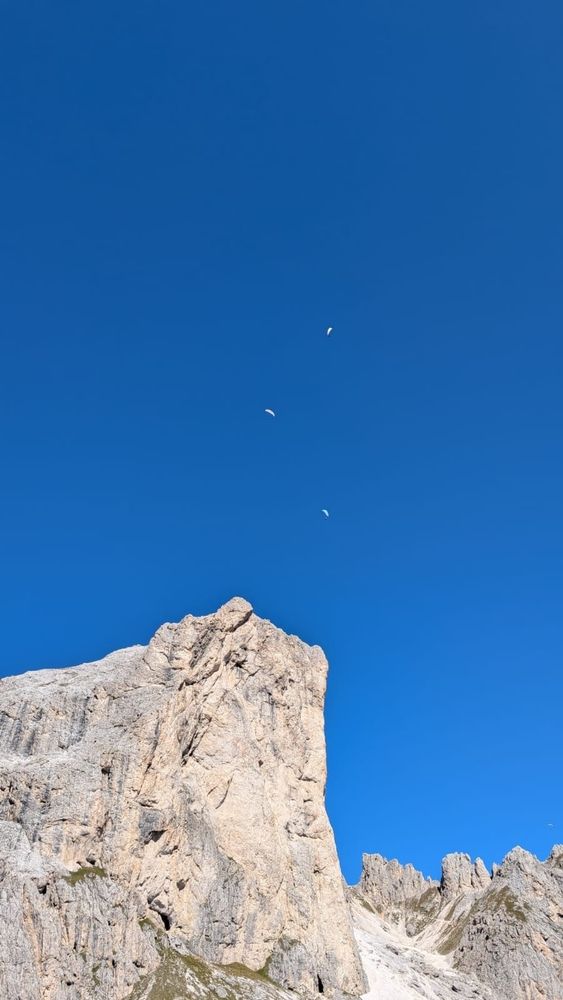 Auf dem Bild ist ein steiler, heller Felsen in einer Gebirgslandschaft zu sehen, der sich markant gegen den wolkenlosen, tiefblauen Himmel abhebt. Über dem Felsen sind drei Gleitschirmflieger zu erkennen, die weit oben am Himmel schweben. Rechts im Hintergrund ragen weitere felsige Gipfel empor.

