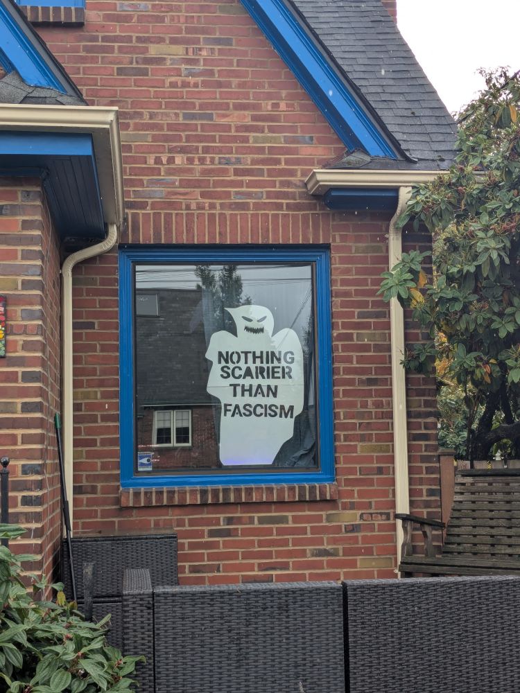 A big window in a brick house. The trim is blue. A big white ghost haunts the window and cut out letters inside it say "Nothing scarier than fascism"