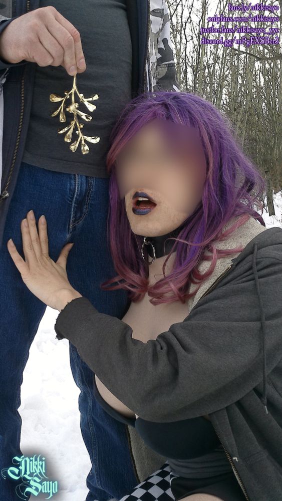 Nikki Sayo is squatting down to bring his face closer to a man's groin while he dangles a mistletoe ornament above the fly