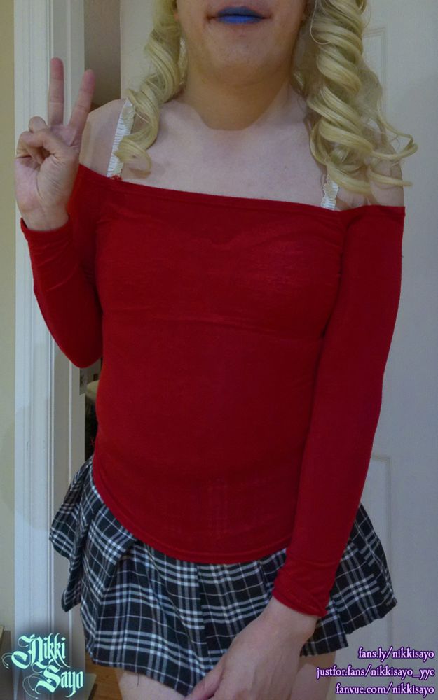Blonde femboy with blue lipstick wearing a red shoulderless top while tugging down the front of their black/white plaid skirt