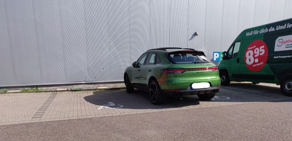 Baumarkt-Parkplatz. Ein grüner Porsche Macan steht quer auf 2 Behinderten-Parkplätzen.