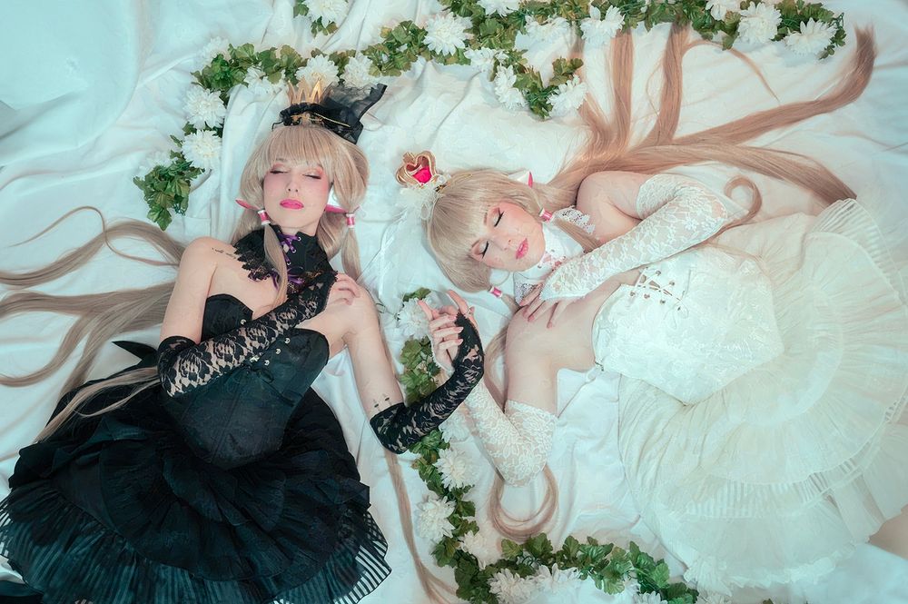 A photo of a Freya and Chii cosplayers laying down on a white sheet with vines of white flowers around them. They are holding hands with their eyes closed and wearing matching dresses in black and white respectively.