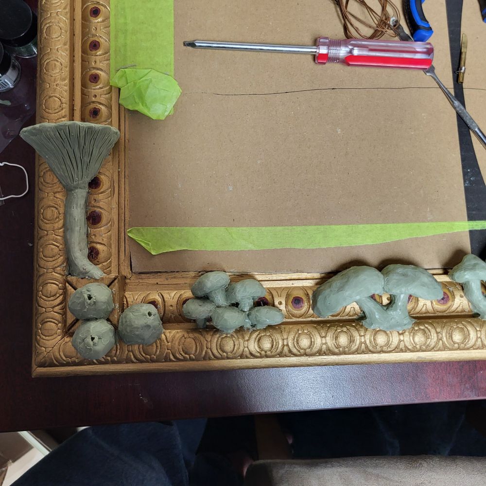 Sculptures of a bunch of bunches of mushrooms growing out of a gold picture frame. Including puffballs, jack o'lanterns and others i can't identify right now.