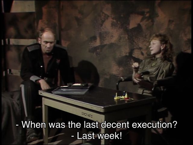 Colin Baker episode. A middle-aged man and woman are sitting in a bizarre room with dull gray, pink and green paint smattered on the walls. On a somewhat industrial looking table, there’s a tray of disappointing food. The man looks sadder than the wall and food combined. The woman is doing some knitting or something. Captions read, “When was the last decent execution?” “Last week!”