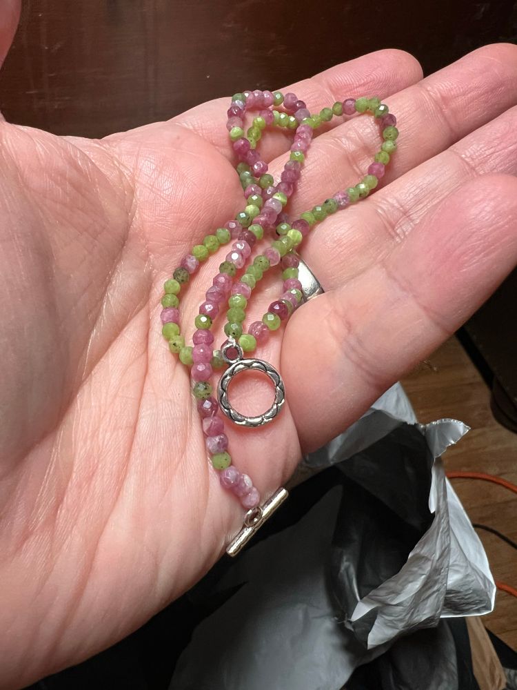 a pale hand holds a necklace of a string of tiny faceted green and pink and pink beads. 