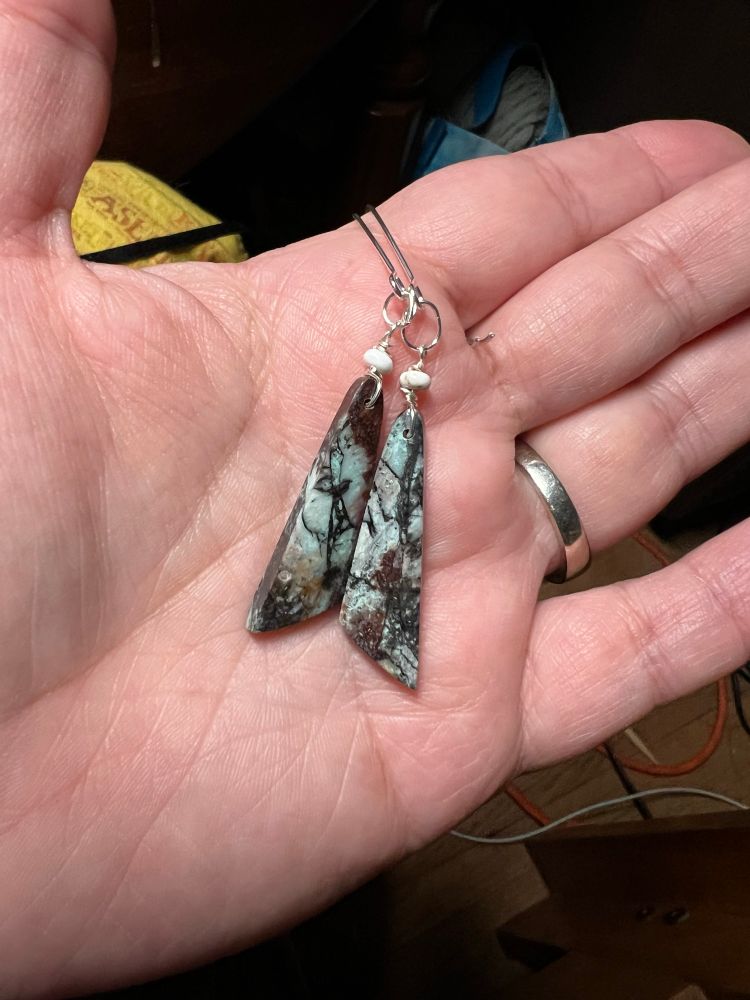 a pale hand holding long silver ear wires with kite shaped stone dangles in a mix of pale green and red webbed with black. 