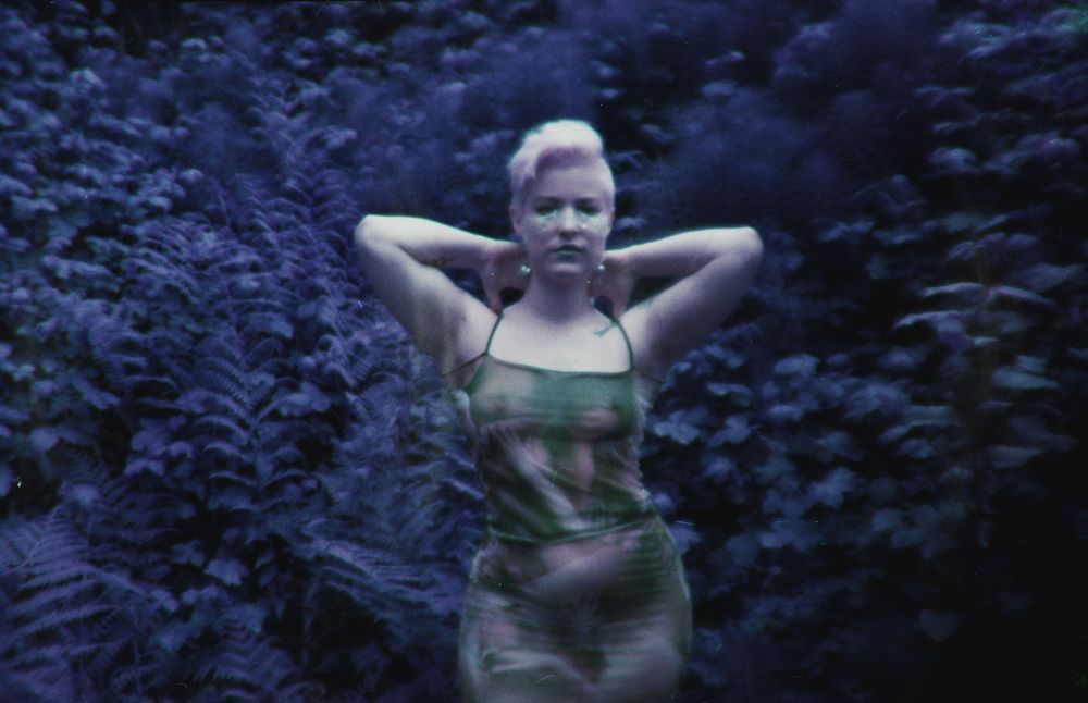 Pepper stands behind a lush scenery of flora and fauna. The photo is tinted in blues and violets because it is taken with infrared film. They wear a sheer shirt and tank top by Four Chambers. They have short platinum hair 