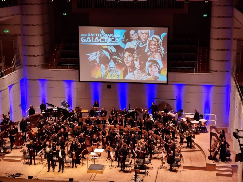 Zu sehen ist ein Orchester und das Titelbild der Serie Kampfstern Galaktika 