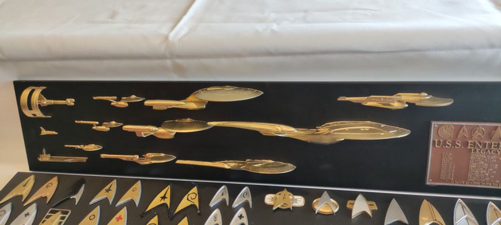 Eine Tafel mit (Zinn?) Modellen aller Enterprises und allen Star Trek Deltas . Zusätzlich ist noch eine Plakette U.S.S. Enterprise zu sehen