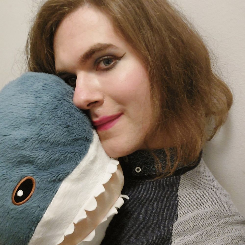 A selfie of me in front of a white wall with a blåhaj covering half my face. I am a white woman with open, slightly curly, shoulder-length brown hair. I am wearing a blue sweater with a grey section from the collar to the shoulders. Underneath, the collar of a blue button-up shirt is visible. I am also wearing make-up consisting of winged eyeliner, pink blush on the cheekbones and pink lipstick