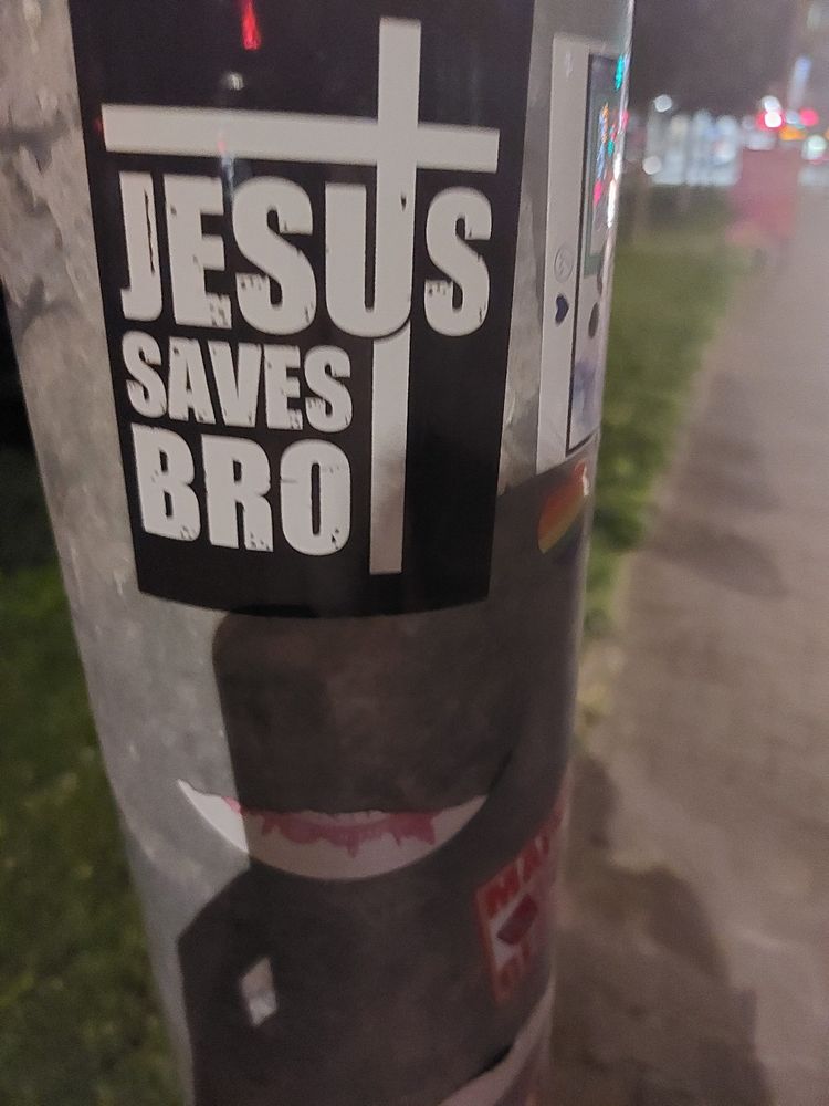 A picture of a sticker on a lamp post. It has white font on a black background. The text reads "Jesus saves bro". There also is a big white cross on the sticker