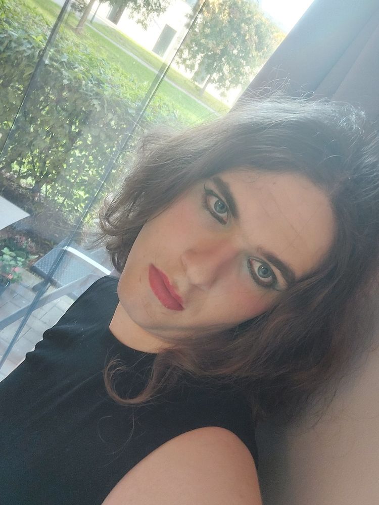 A selfie of me wearing a black dress. I am a 20 something white woman. I am wearing make-up, which consists of winged eyeliner, blue eyeshadow with glitter, red lipstick, and pink blush that covers the entirety of my cheekbones and also goes over my nose. In the top left of the picture, there is a window through which the corner of a table and a chair standing on a stone floor are visible. Behind the stone floor, there is a hedge