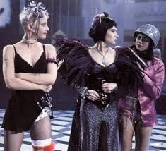A pic from the movie Tank Girl. On the left is Tank Girl wearing a short black tank dress and ribbons in her short blonde hair. In the middle is the Madam wearing a black dress with black feathered shoulder pieces. On the right Jet is holding a gun on the madam, she is wearing a shiny pink jacket, shorts, and a sparkly silver helmet.
