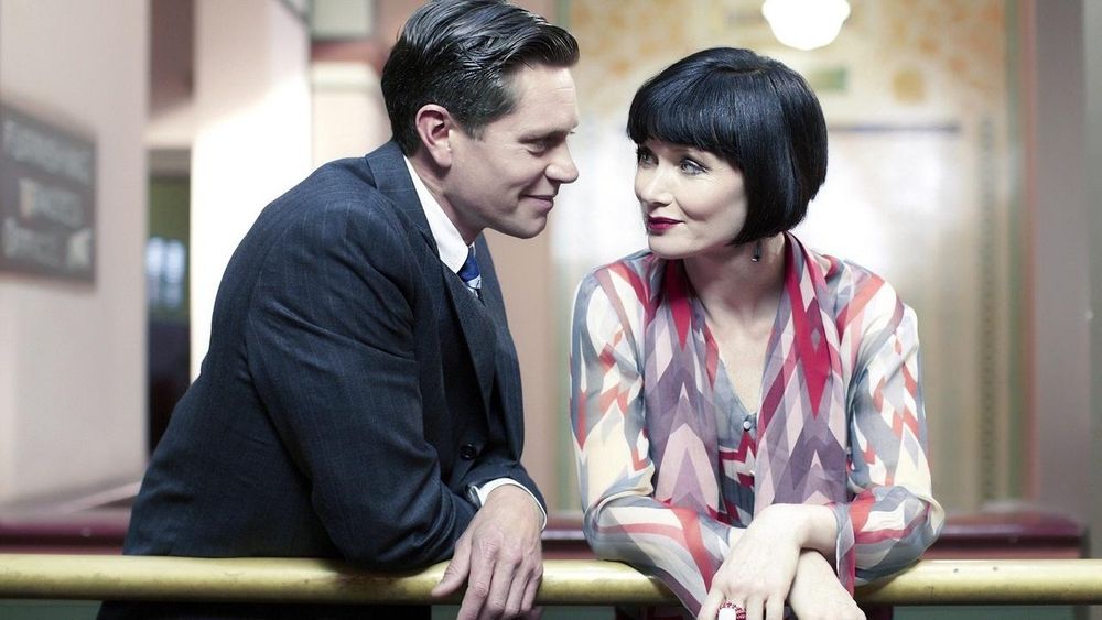 Jack in a tailored suit doing The Lean and smirking at Phryne, who is smiling right back.