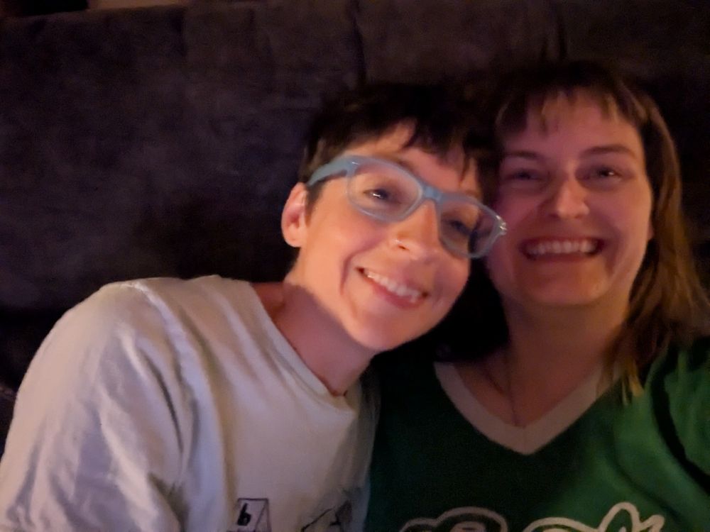 My wife and I at home on the couch wearing comfy clothes. We're snuggling and laughing while trying to take a selfie. 