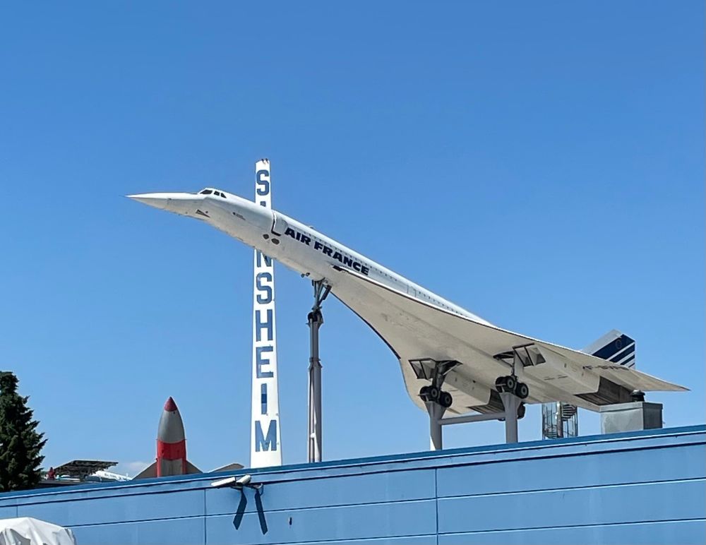 Concorde von schräg unten fotografiert im Museum Sinsheim. Man sieht das Fahrwerk und im Hintergrund vor dem blauen Himmel den senkrechten Schriftzug „Sinsheim“
