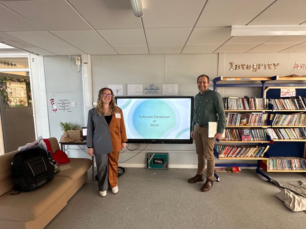 A woman and a man standing at either side of an interactive panel in a school library. The panel is showing a PowerPoint slide with ‘Software Developer At Work’ on it. 