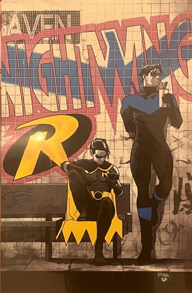 Nightwing and Robin are sitting at a bus or subway stop. Robin is on his phone, nightwing is sipping a soda. Graffiti with their logos is depicted on the wall behind them.