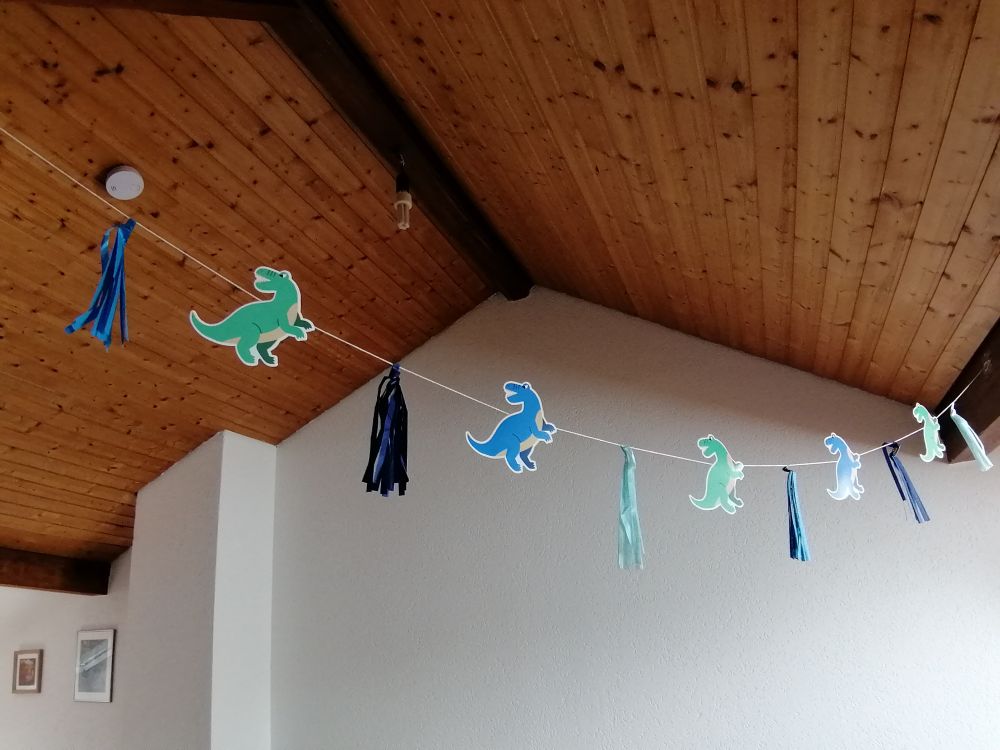 Bunting with dinosaurs hanging from a wood ceiling. White wall as background and a couple of frames.

Cenefa con dinosaurios colgando del techo de madera. Una pared blanca de fondo con unos cuadros.