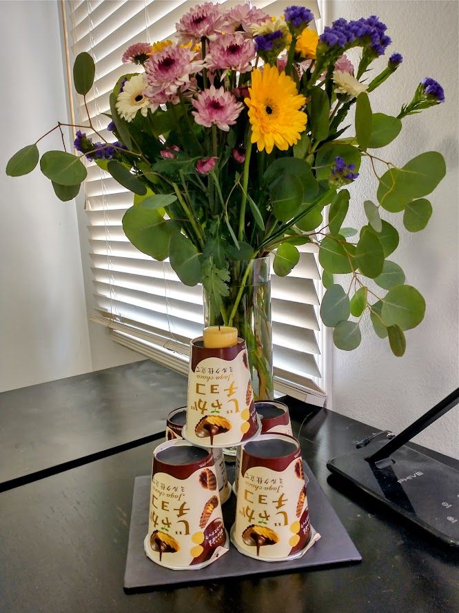 A bouquet of flowers and decorative leaves in a glass vase, with a stack of Japanese chocolate covered potato chip cups topped with a candle, serving as a birthday cake.