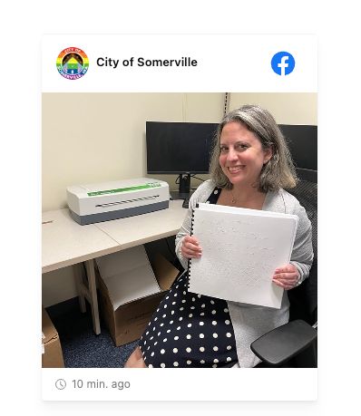 Post to Facebook by the official City of Somerville social media account. The only text in the post preview appears to be "10 min. ago." The post is a photo of a smiling white woman with grey hair holding some Braille-embossed papers in front of her. She is in an office setting next to a desk. There is a computer in the background behind her, and the computer is next to a IRIE BrailleTrac 120 desktop Braille embosser which is in white, green, and grey plastic.