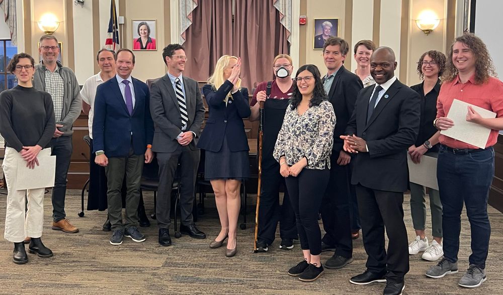 Thirteen people (mostly white people, one black man) in a formal building (Somerville City Hall). Many are holding certificates and smiling. 