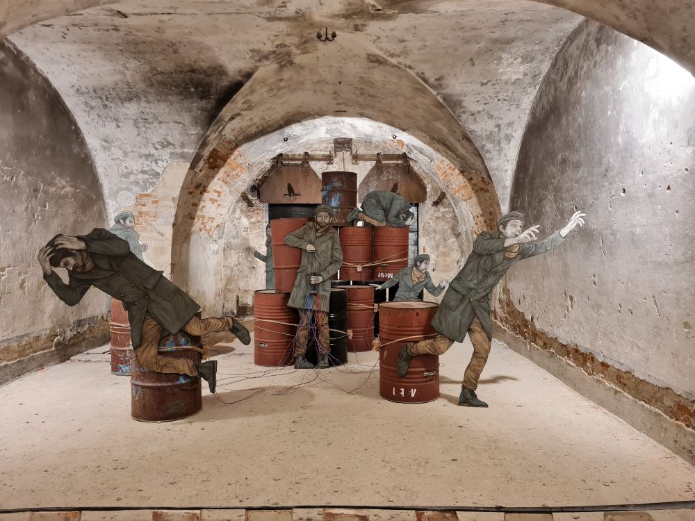In einem alten, gewölbten Kellerraum stehen rostige Ölfässer, umgeben von lebensgroßen, flachen Figuren in historischer Kleidung. Die Figuren wirken wie Soldaten im Schockmoment – einige ducken sich, andere fliehen oder schreien. Bunte Kabel verbinden die Fässer, was an eine Explosion oder ein gefährliches Szenario erinnert. Die Szene vermittelt Chaos, Angst und Flucht, eingebettet in die raue, geschichtsträchtige Atmosphäre des Ortes.
